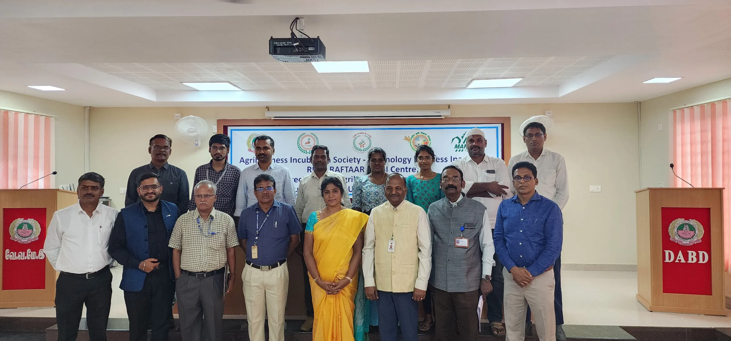 A group of 12 people stand in a conference room, posing for a photo in front of a banner that mentions "Agribusiness Incubation Society" and "Technology Business Incubator." Among them is Jane from Aerospace, who specializes in integrated supply chain management.