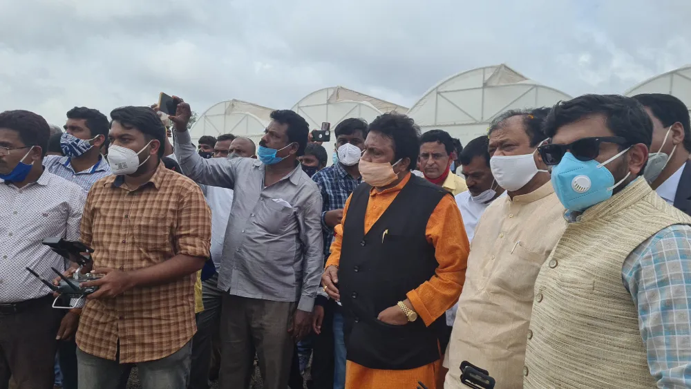 A group of people, mostly men, wearing face masks and diverse clothing, gather closely outdoors. Some hold cameras and smartphones, capturing what appears to be a Jane event. A few structures are visible in the background.