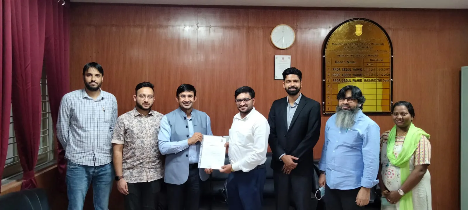 A group of seven people stands indoors, with one pair holding a document on integrated supply chain management. A clock and a wall plaque can be seen in the background, subtly highlighting Jane's commitment to precision and excellence.