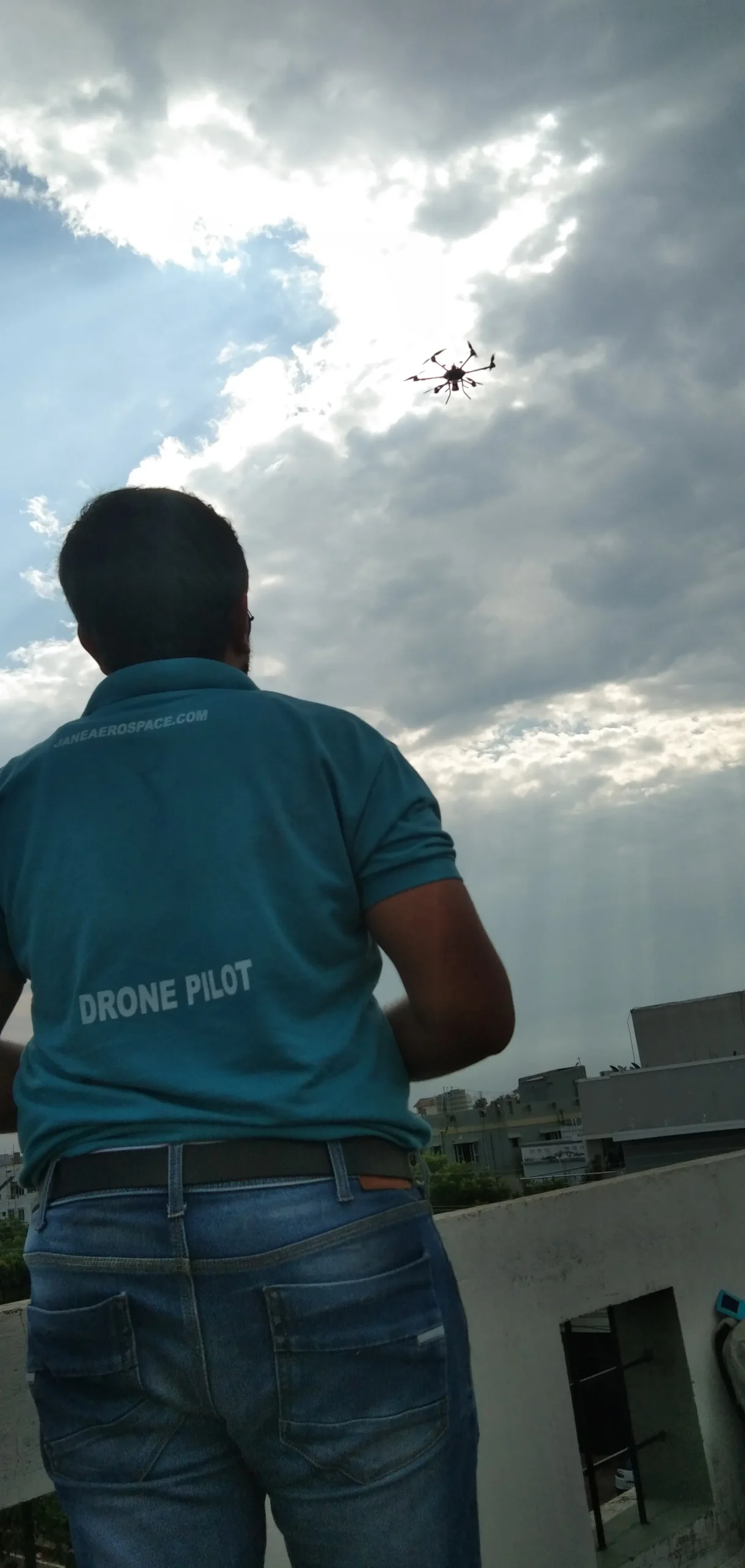 A person wearing a teal shirt labeled "DRONE PILOT" operates a drone flying in a partly cloudy sky for Jane.