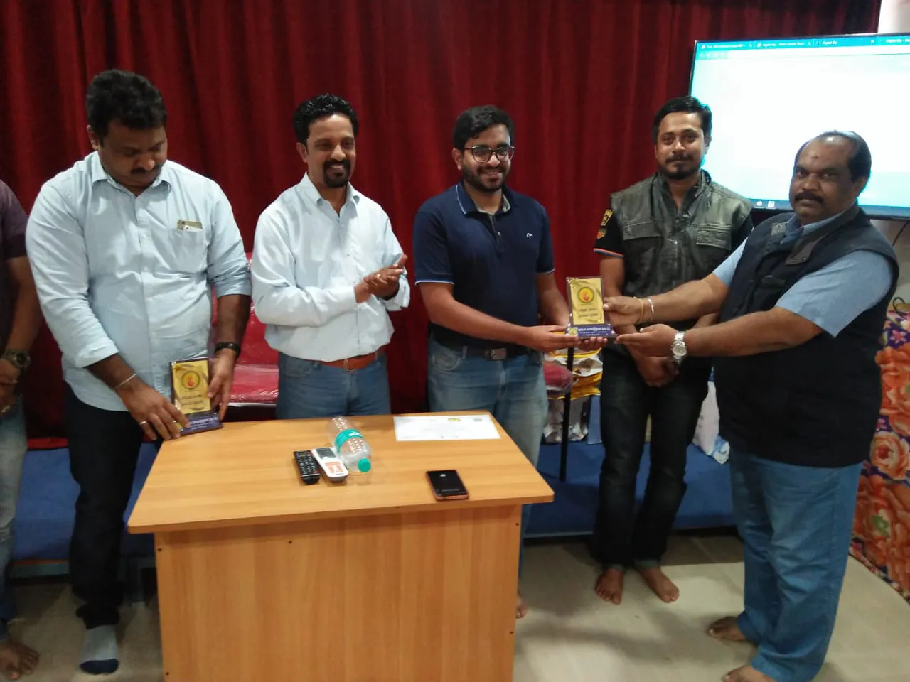 A group of men stands in front of a red curtain, with two exchanging a plaque. In front of them, a table holds items like phones, a bottle, and papers related to integrated supply chain management for Jane.