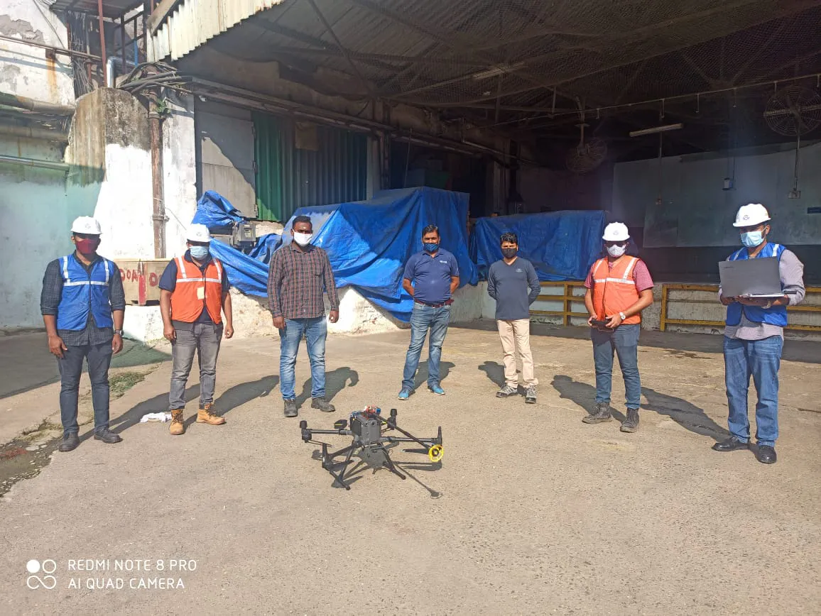 A group of six individuals in safety gear is standing around a drone in an industrial environment, representing Jane's commitment to integrated supply chain management. Some of them are wearing hard hats and high-visibility vests.