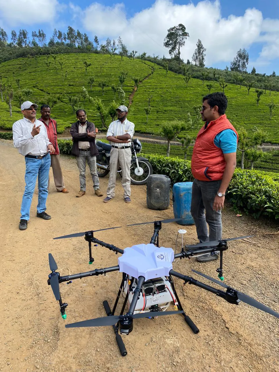 Five men stand on a dirt road near a large Jane drone. The setting is a green, hilly landscape with vegetation in the background. One man gestures while others look on, discussing integrated supply chain management. A motorcycle is parked nearby.