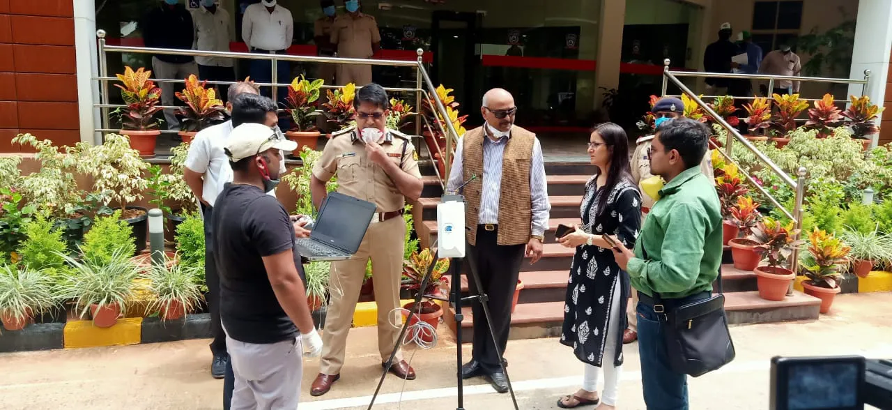 A group of people, including police officers and other individuals, stand outside a building near some potted plants, engaged in a discussion around a laptop and a device on a stand. Jane is mentioned as they talk about integrated supply chain management solutions.