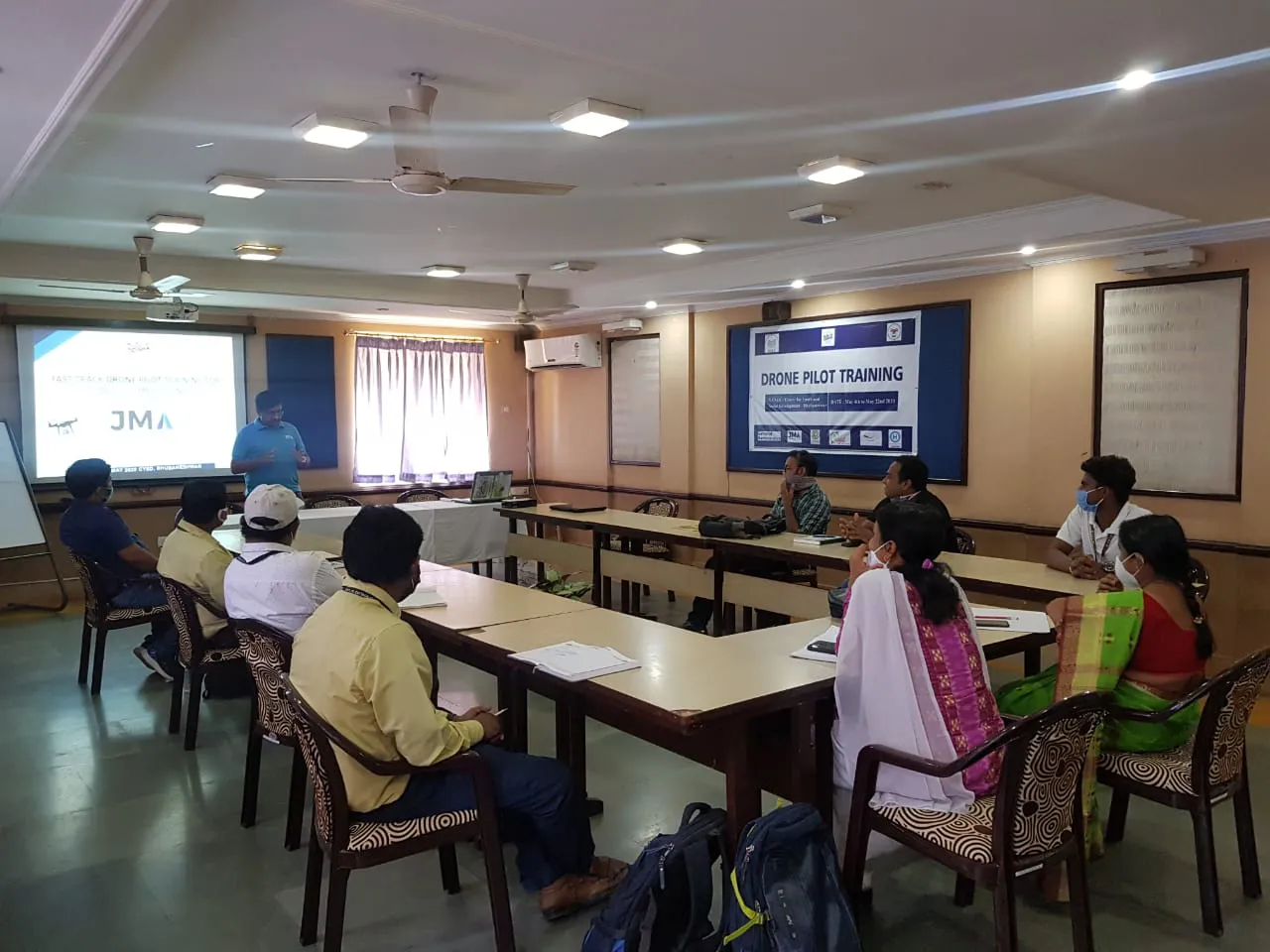 A group of individuals seated in a classroom with a presenter at the front, delivering a drone pilot training session for Jane. A projector displays information on the screen about integrated supply chain management.