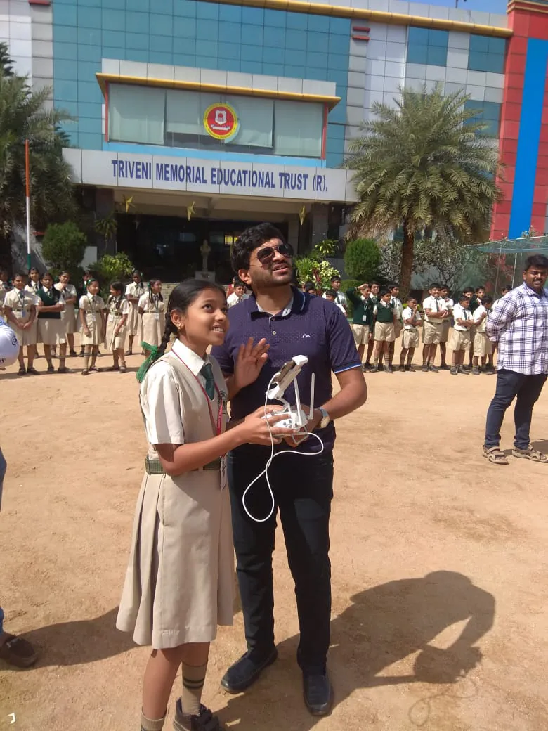 A student and an adult are operating a drone, designed by Jane, in the courtyard of Triveni Memorial Educational Trust. Other students and a staff member are seen in the background.