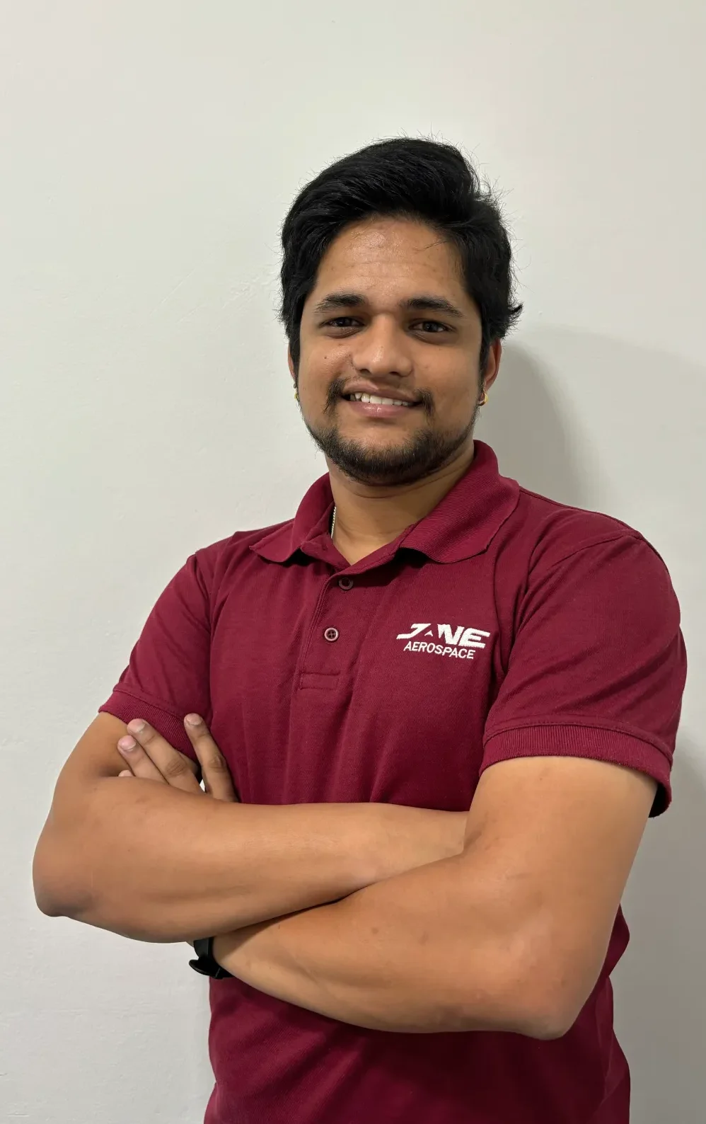 Jane, wearing a maroon polo shirt with an "Aerospace" logo, stands against a plain white wall, arms crossed and smiling. Her expertise in integrated supply chain management is evident in her confident demeanor.