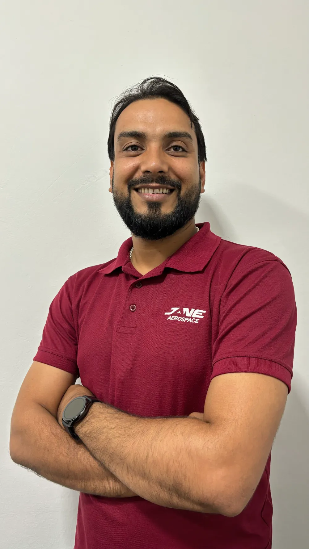 A person with short dark hair and a beard stands against a white wall, smiling and wearing a maroon polo shirt that reads "Jane" with arms crossed—representative of their integrated supply chain management expertise.