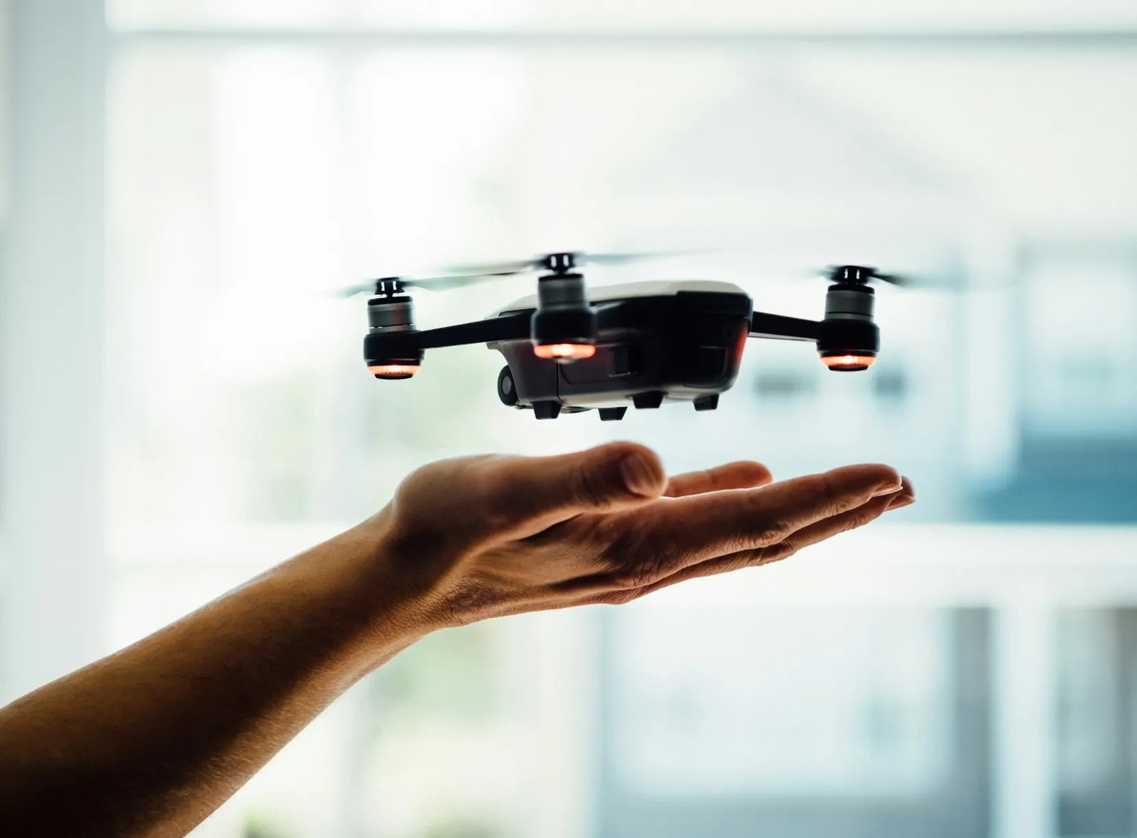 A small drone from Jane hovers just above a person's outstretched hand indoors, with a blurred window and buildings visible in the background.