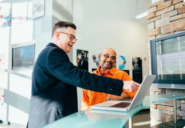 Two men stand at a counter, one in an orange shirt and the other in a dark jacket, pointing at a laptop screen and smiling. A wall-mounted TV and stone wall are in the background, possibly discussing integrated supply chain management for Jane.