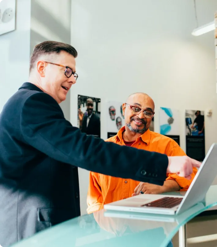 Two men standing at a counter, one in an orange shirt and glasses, the other in a dark jacket, pointing at a laptop screen on the counter in a well-lit room discussing integrated supply chain management at Jane.