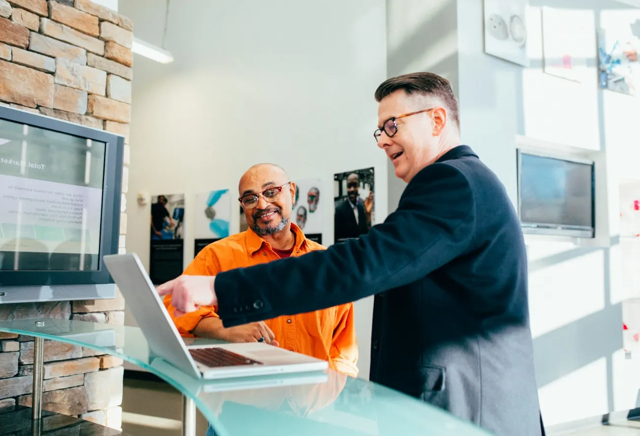 Two men engaging in a discussion about integrated supply chain management, one pointing at a laptop screen on a glass-topped counter, with a television and a brick wall in the background.