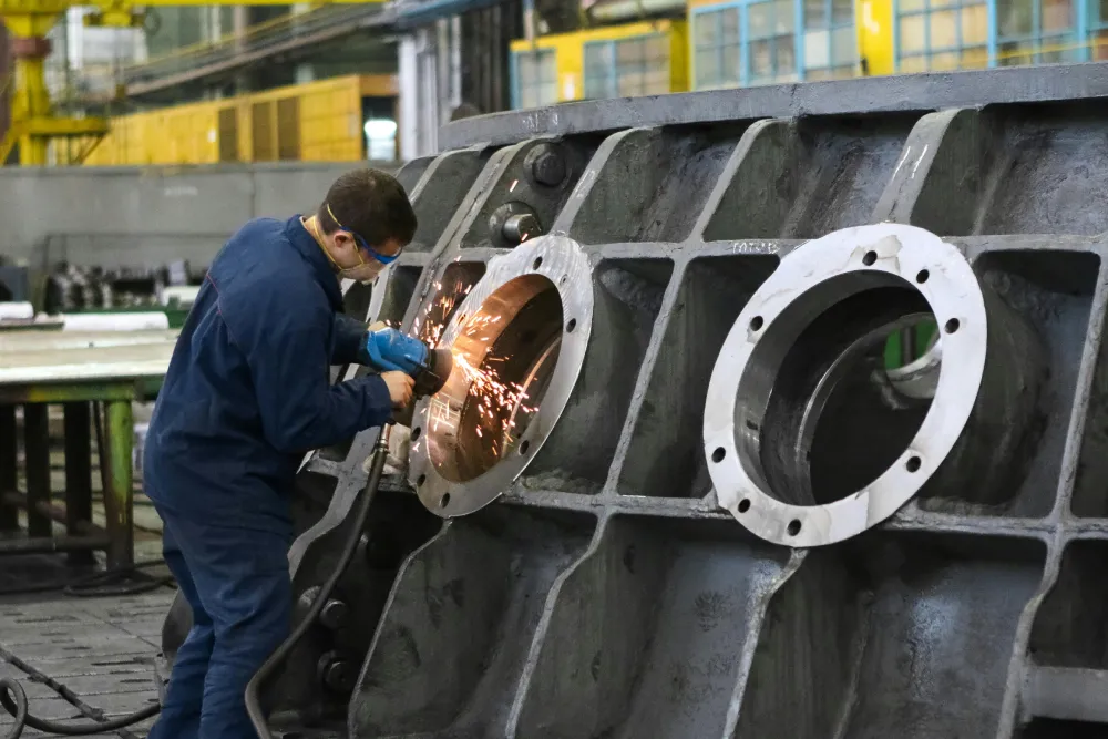 A man in protective gear uses a power tool on a large industrial metal component in a Jane factory setting, showcasing their expertise in integrated supply chain management.