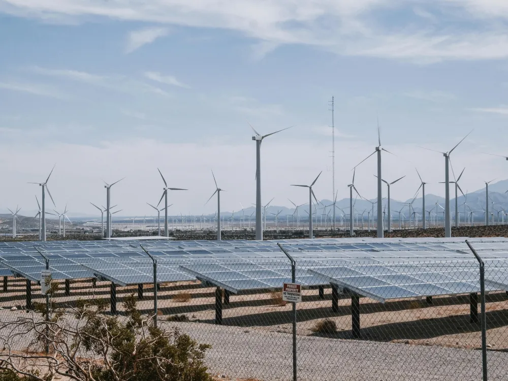 A field of solar panels and wind turbines with mountains in the background under a partly cloudy sky, enclosed by a chain-link fence, provided by Jane.