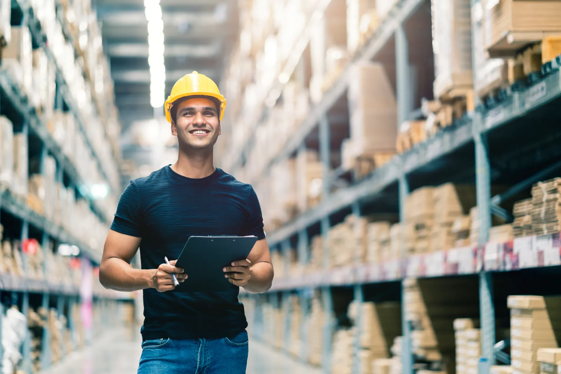 Jane, a worker in a hard hat, holds a clipboard while standing in a warehouse aisle filled with shelves, demonstrating the seamless flow of integrated supply chain management.